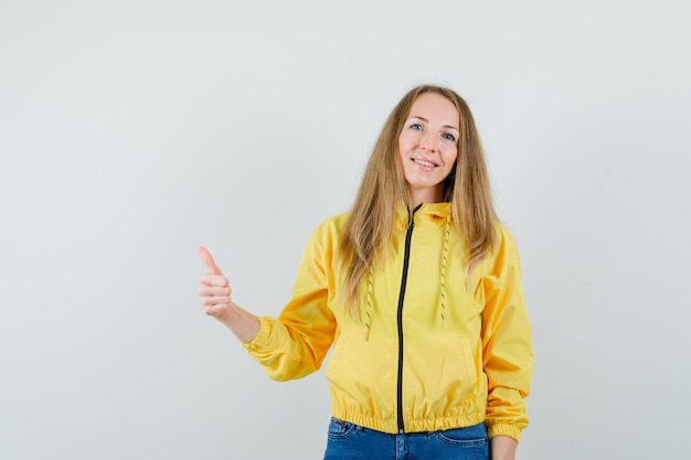 Mujer joven mostrando el pulgar hacia arriba en chaqueta de bombardero amarilla y jean azul y mirando sorprendido. vista frontal.