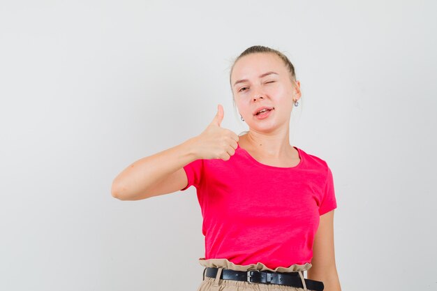 Mujer joven mostrando el pulgar hacia arriba, guiñando un ojo en la vista frontal de la camiseta y los pantalones