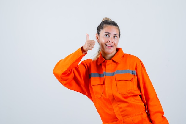 Foto gratuita mujer joven mostrando el pulgar hacia arriba mientras sonríe en uniforme de trabajador y parece divertido, vista frontal.