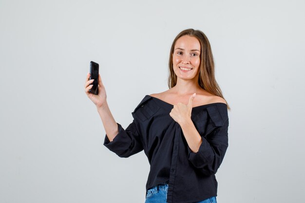 Mujer joven mostrando el pulgar hacia arriba mientras sostiene el teléfono inteligente en camisa, pantalones cortos y mirando alegre