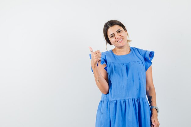 Mujer joven mostrando el pulgar hacia arriba en vestido azul y mirando feliz