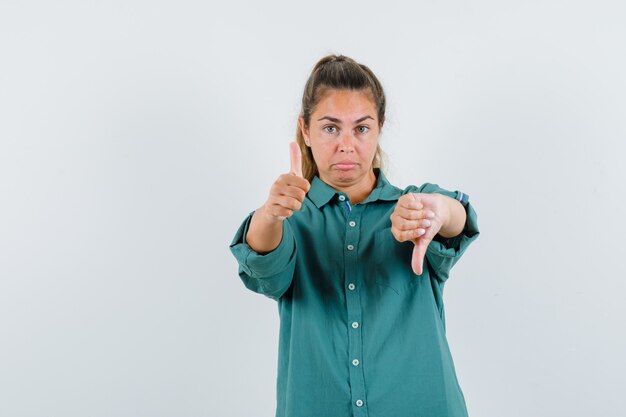 Mujer joven mostrando los pulgares hacia arriba y hacia abajo con ambas manos, haciendo muecas en blusa verde y luciendo lindo