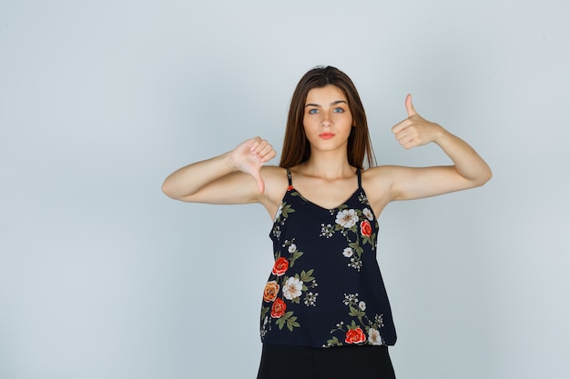 Mujer joven mostrando los pulgares hacia arriba y hacia abajo en la parte superior floral y mirando melancólica