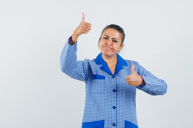 Mujer joven mostrando los pulgares para arriba con ambas manos en camisa de pijama de cuadros azules y mirando bonita, vista frontal.