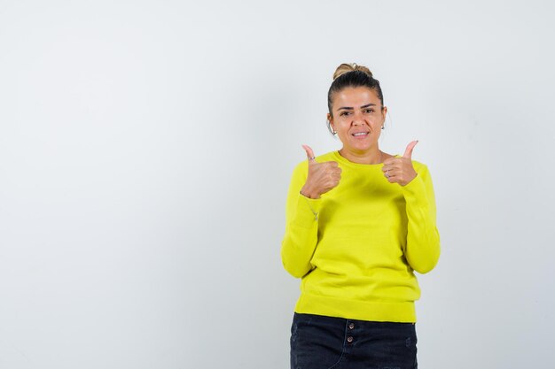 Mujer joven mostrando los pulgares para arriba con ambas manos en suéter amarillo y pantalón negro y mirando feliz