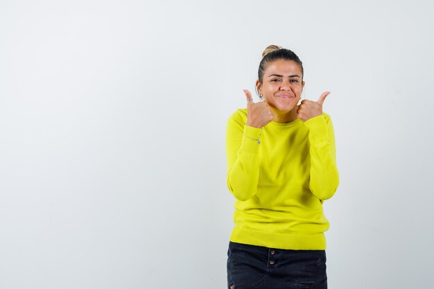 Mujer joven mostrando los pulgares para arriba con ambas manos en suéter amarillo y pantalón negro y mirando feliz