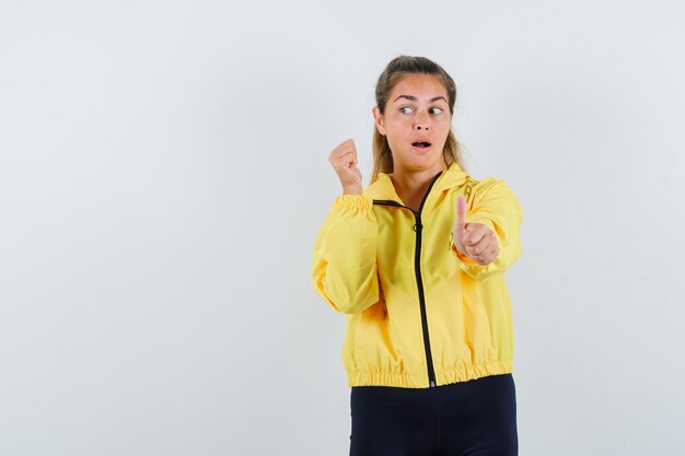 Mujer joven mostrando los pulgares hacia arriba y apretando el puño mientras mira a la izquierda en una chaqueta de bombardero amarilla y pantalones negros y se ve linda