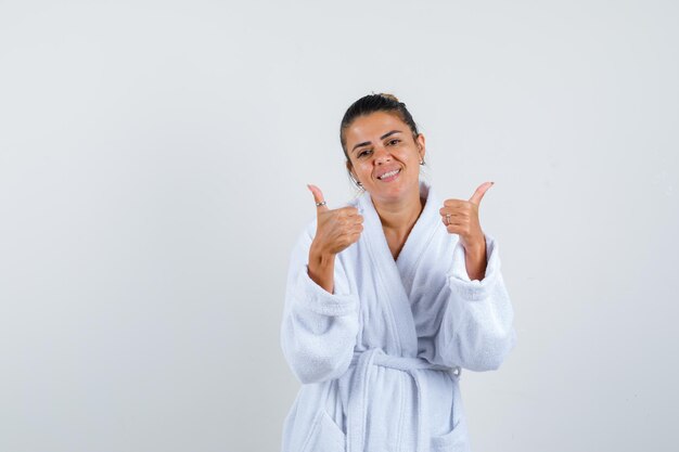 Mujer joven mostrando los pulgares para arriba en bata de baño y mirando feliz