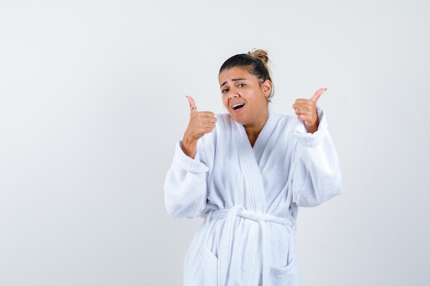 Mujer joven mostrando los pulgares para arriba en bata de baño y mirando feliz
