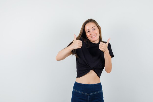 Mujer joven mostrando los pulgares para arriba en blusa negra, pantalones y mirando feliz