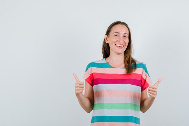 Mujer joven mostrando los pulgares para arriba en camiseta y mirando feliz, vista frontal.
