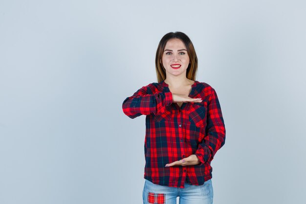 Mujer joven mostrando signo de gran tamaño en camisa a cuadros y mirando alegre. vista frontal.