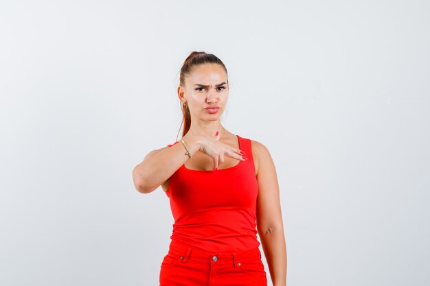 Mujer joven mostrando signo de mano de pistola en camiseta roja sin mangas, pantalones y mirando confiado, vista frontal.