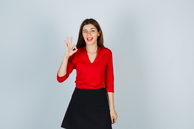 Mujer joven mostrando el signo de ok, sacando la lengua en blusa roja