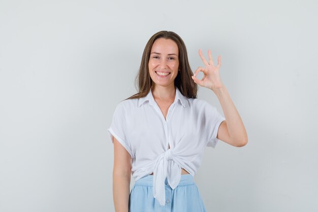 Mujer joven mostrando el signo de ok y sonriendo con blusa blanca y falda azul claro y mirando alegre