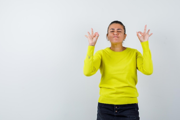 Mujer joven mostrando signos de ok, cerrando los ojos con suéter amarillo y pantalón negro y mirando tranquilo