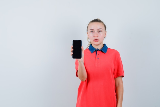 Mujer joven mostrando teléfono móvil en camiseta y mirando desconcertado