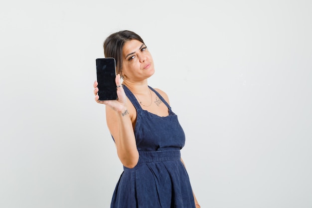 Mujer joven mostrando teléfono móvil en vestido y mirando serio