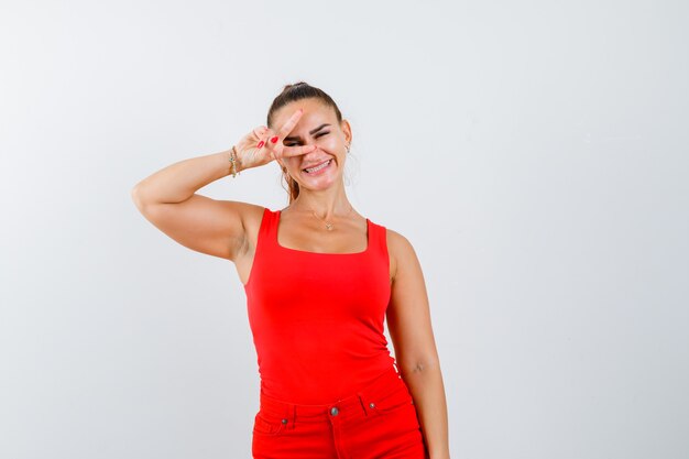 Mujer joven mostrando V-sign en camiseta roja, pantalones y mirando alegre, vista frontal.