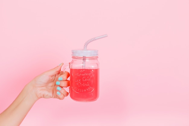 Mujer joven mostrando un vaso con jugo de bayas recién exprimido, que hizo para el almuerzo con la familia. Foto de taza grande con limonada sabrosa brillante para fiesta de cumpleaños aislado sobre fondo rosa