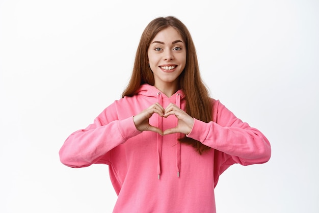 La mujer joven muestra el signo del corazón y sonríe Me gusta el gesto de amarte expresar emociones románticas de pie sobre fondo blanco