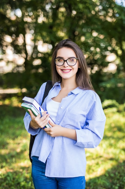 Mujer joven muy atractiva con libros de pie y sonriendo en el parque