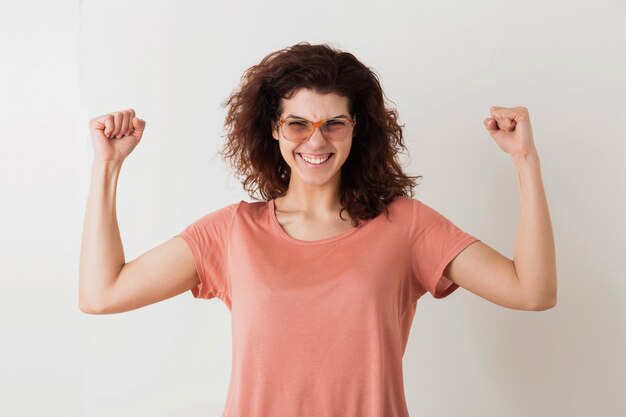 Mujer joven muy elegante con gafas sosteniendo las manos en alto en gesto de victoria, músculos fuertes, emocional, ganador, cabello rizado, sonriente, emoción positiva, feliz, aislado, camiseta rosa, estilo hipster