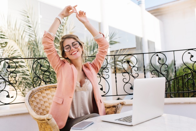 Mujer joven muy feliz disfrutando del tiempo, relajándose en la acogedora cafetería al aire libre. Portátil plateado sobre mesa y smartphone. Lleva gafas elegantes, chaqueta rosa, blusa de encaje beige, relojes blancos.