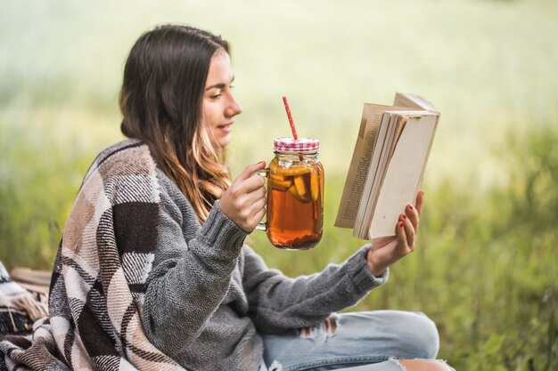 Mujer joven en la naturaleza con una bebida en la mano leyendo un libro