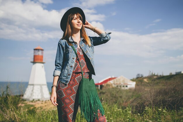 Mujer joven en la naturaleza, faro, traje bohemio, chaqueta vaquera, sombrero negro, sonriente, feliz, verano, accesorios elegantes