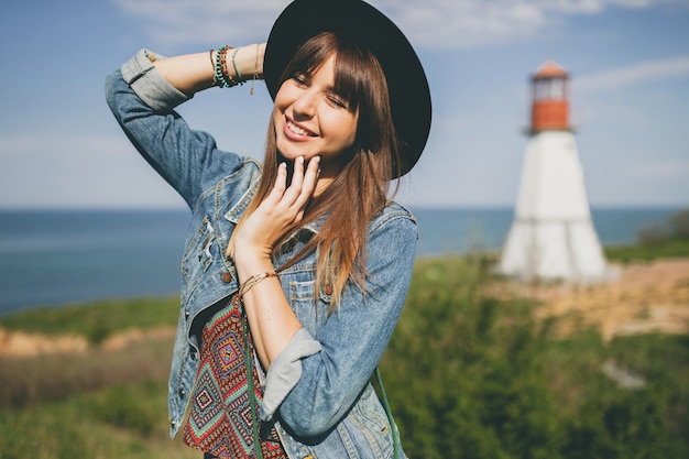 Mujer joven en la naturaleza, faro, traje bohemio, chaqueta vaquera, sombrero negro, sonriente, feliz, verano, accesorios elegantes