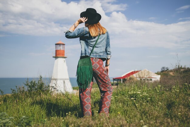 Mujer joven en la naturaleza, faro, traje bohemio, chaqueta vaquera, sombrero negro, verano, accesorios elegantes