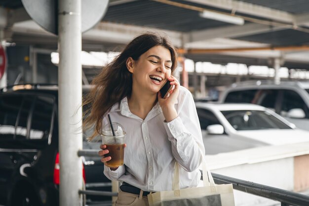 Mujer joven de negocios con café hablando alegremente por teléfono en el estacionamiento