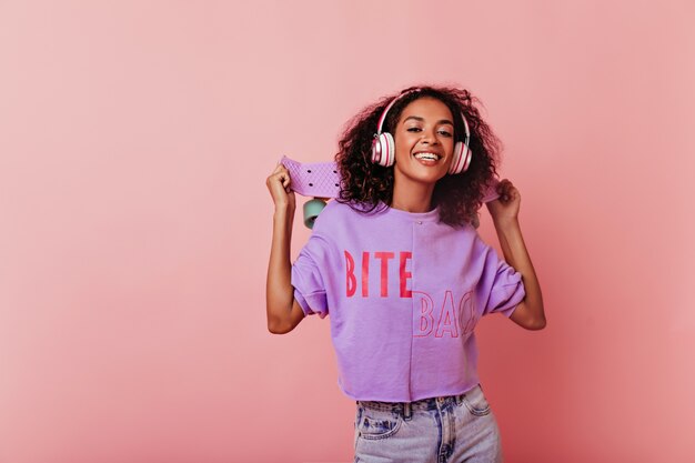 mujer joven negra de pie en pose segura con patineta. Impresionante chica africana rizada en auriculares aislados en pastel en estudio.