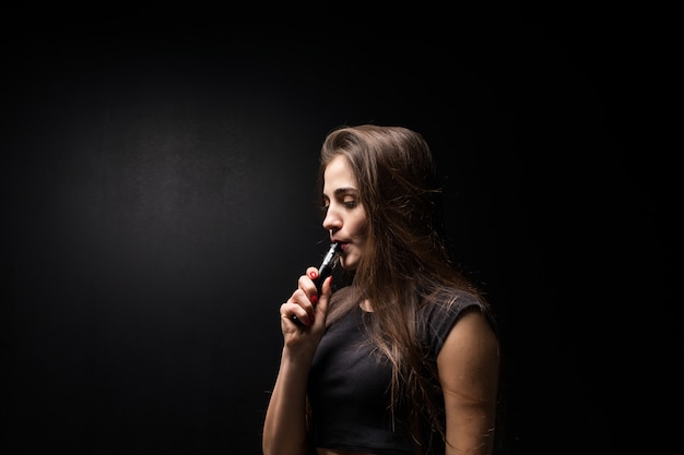 Foto gratuita mujer joven en negro fuma un cigarrillo electrónico en la pared oscura