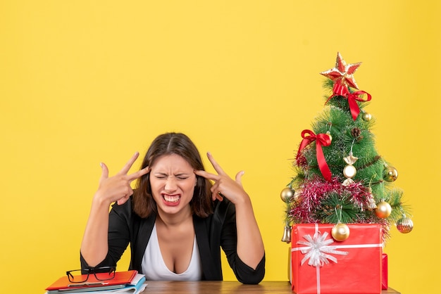 Mujer joven nerviosa y emocional enojada sentada en una mesa cerca del árbol de Navidad decorado en la oficina en amarillo