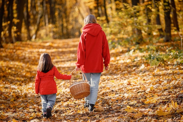 Mujer joven con niña caminando en el bosque de otoño. Mujer rubia juega con su hija y sostiene una canasta. Madre e hija con jeans y chaquetas rojas.