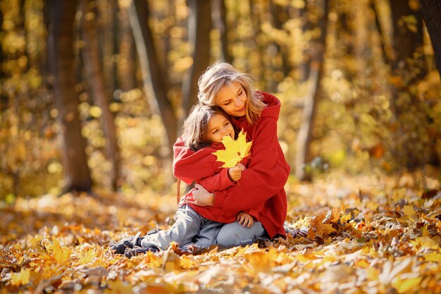 Mujer joven con niña sentada sobre una manta en el bosque de otoño. Mujer rubia juega con su hija. Madre e hija con jeans y chaquetas rojas.