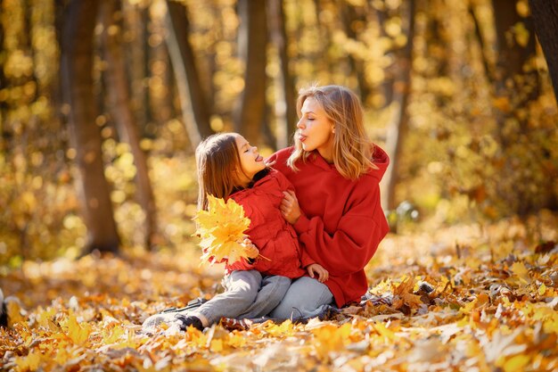 Mujer joven con niña sentada sobre una manta en el bosque de otoño. Mujer rubia juega con su hija. Madre e hija con jeans y chaquetas rojas.