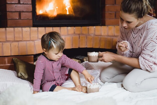 Mujer joven con un niño junto a la chimenea. Madre e hijo beben cacao con marshmello cerca de la chimenea.