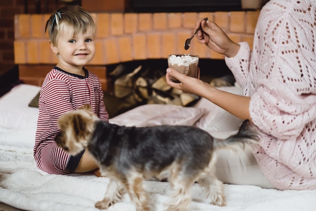 Mujer joven con un niño junto a la chimenea. Madre e hijo beben cacao con marshmello cerca de la chimenea.