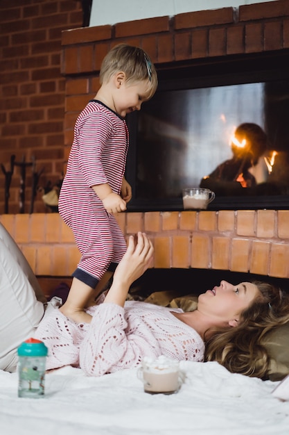 Mujer joven con un niño. Mamá e hijo están jugando, divirtiéndose cerca de la chimenea.