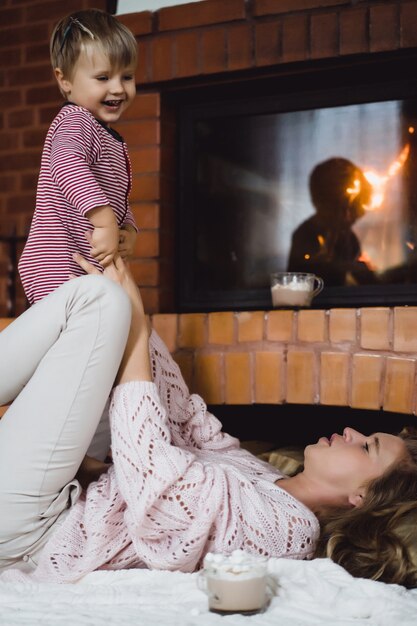 Mujer joven con un niño. Mamá e hijo están jugando, divirtiéndose cerca de la chimenea.