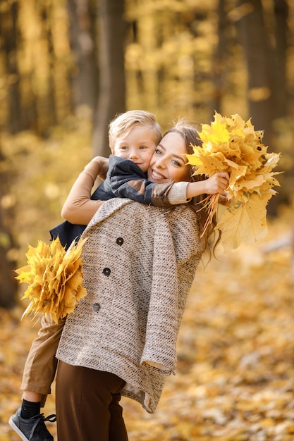 Mujer joven y niño pequeño en el bosque de otoño. Mujer sosteniendo a su hijo en sus manos. Niño usando ropa de moda y sosteniendo hojas amarillas.