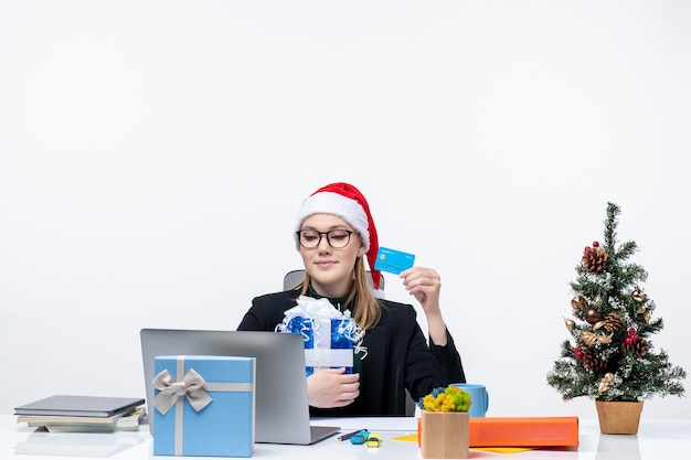 Mujer joven ocupada con sombrero de santa claus y gafas sentado en una mesa con regalo de Navidad y tarjeta bancaria sobre fondo blanco.