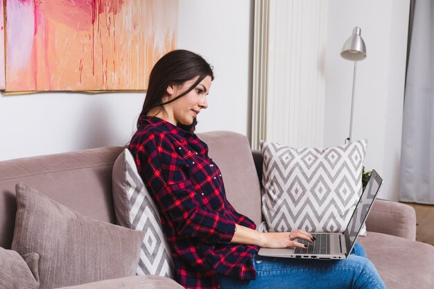 Mujer joven ocupada en usar la computadora portátil que se sienta en el sofá