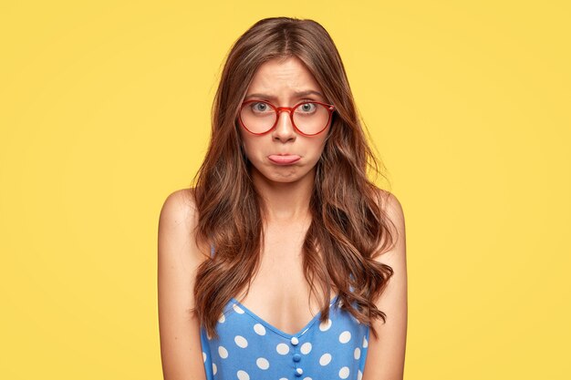 Mujer joven ofendida con gafas posando contra la pared amarilla