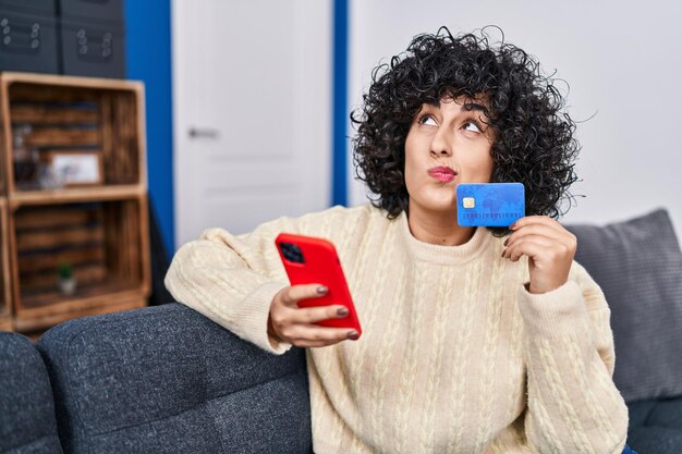 Mujer joven de oriente medio usando teléfono inteligente y tarjeta de crédito sentada en el sofá en casa