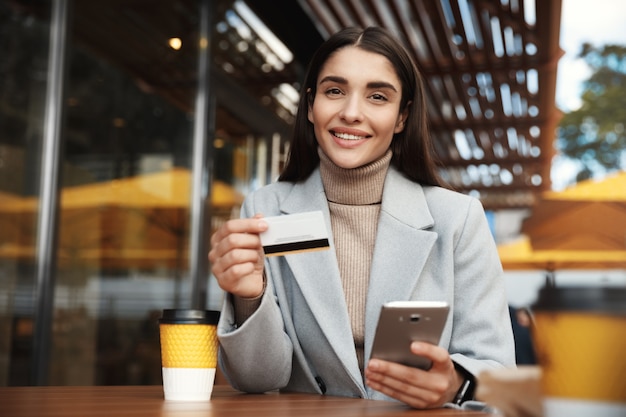 Mujer joven pagando en línea, con tarjeta de crédito y teléfono móvil mientras está sentado en una cafetería.