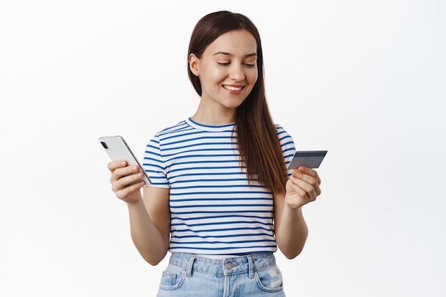 Mujer joven pagando con tarjeta de crédito y teléfono móvil, sonriendo y mirando relajado, compra algo en la tienda de Internet, comprando en la aplicación de teléfono inteligente, de pie sobre una pared blanca.